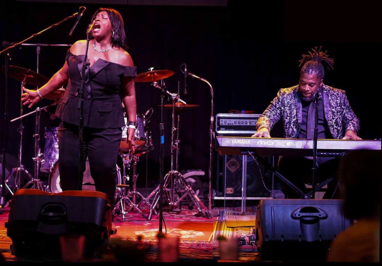 A woman sings at a microphone and a man plays a keyboard on a stage with a drum set in the background.