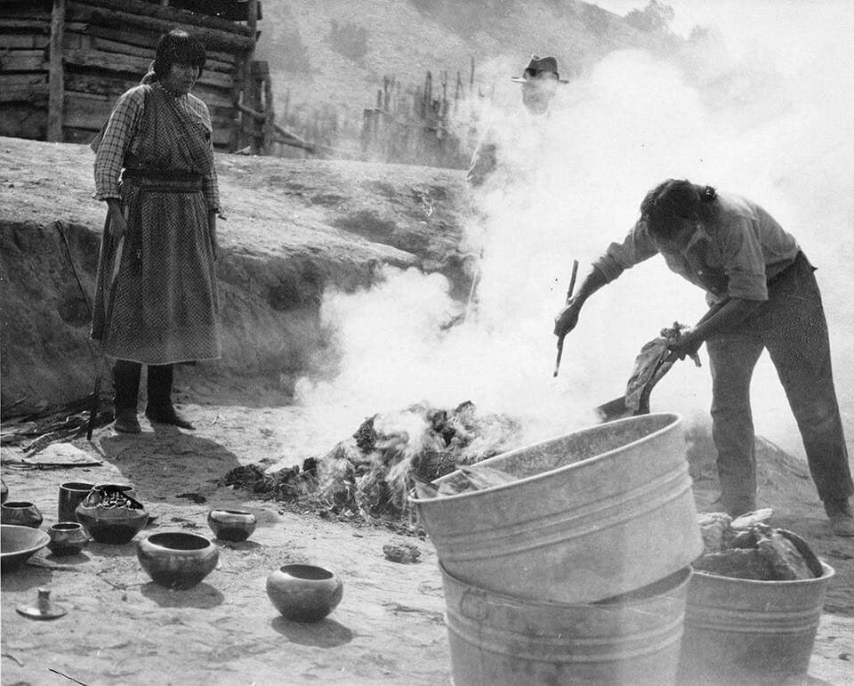 A black-and-white photo of a man agitating a smoking pile of ash with a poker. In the foreground there are steel pales and glistening works of pottery resting on the ground. A woman stands and watches the man work. 