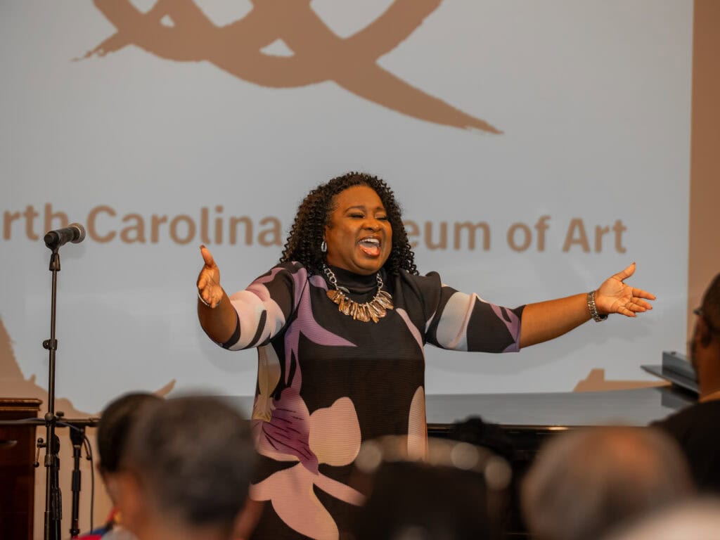 Tiffany Bennett-Cornelous stands with mouth open and arms spread wide next to a microphone and in front of a group of seated people.