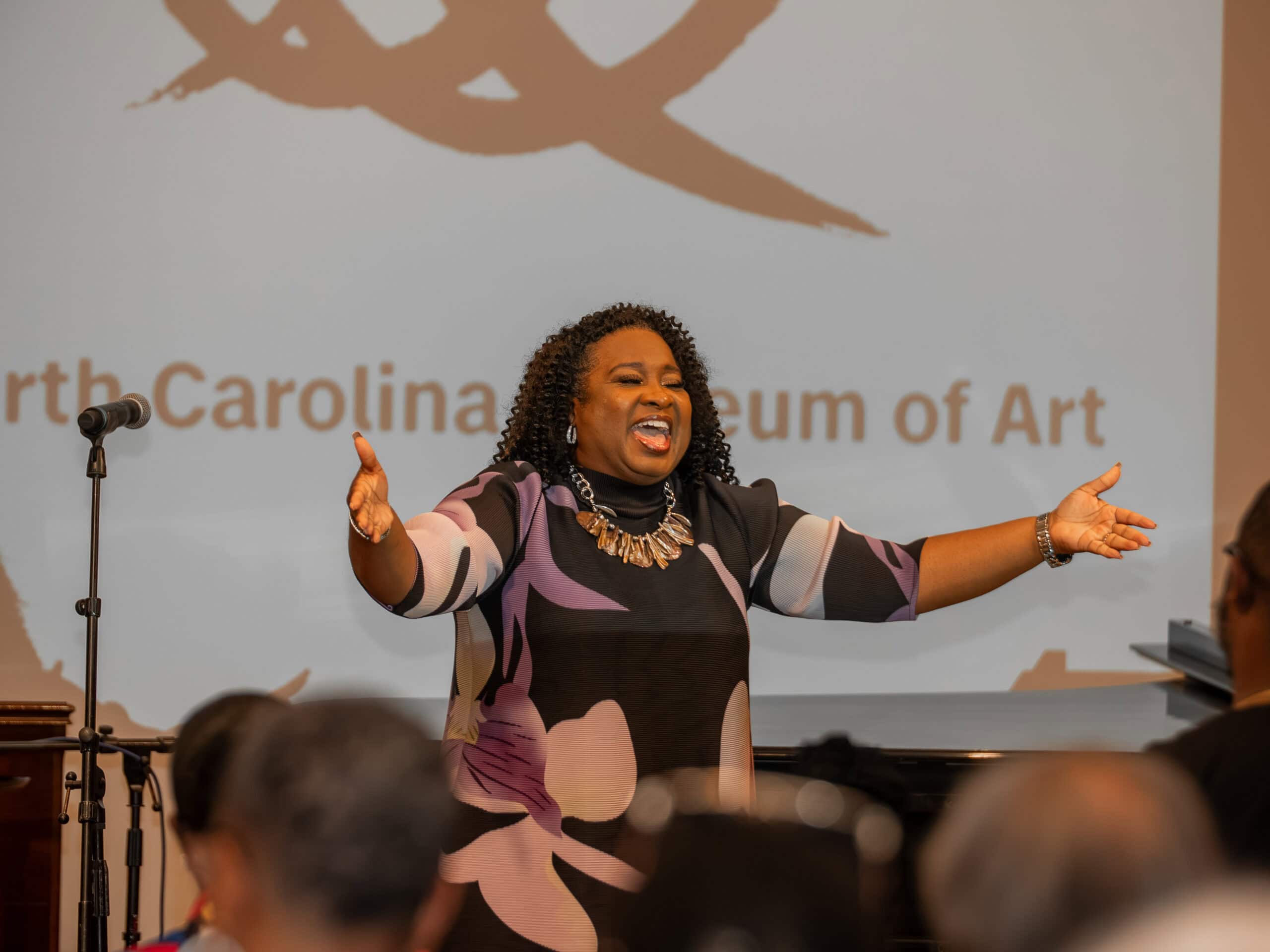 Tiffany Bennett-Cornelous stands with mouth open and arms spread wide next to a microphone and in front of a group of seated people.