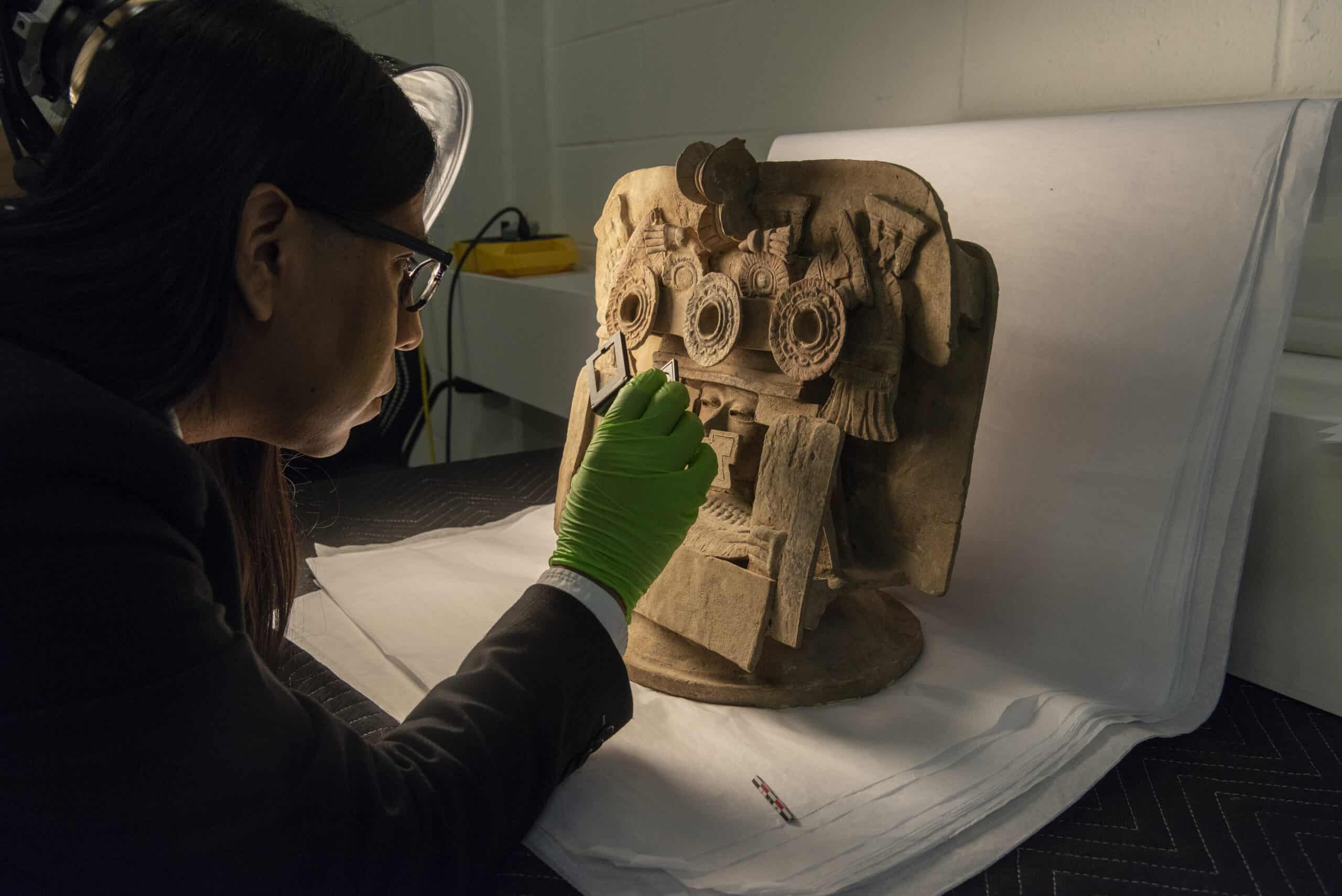 Ángel Gonzalez examines a ceramic sculpture placed on sheets of white paper using a strong light and magnifying glass.