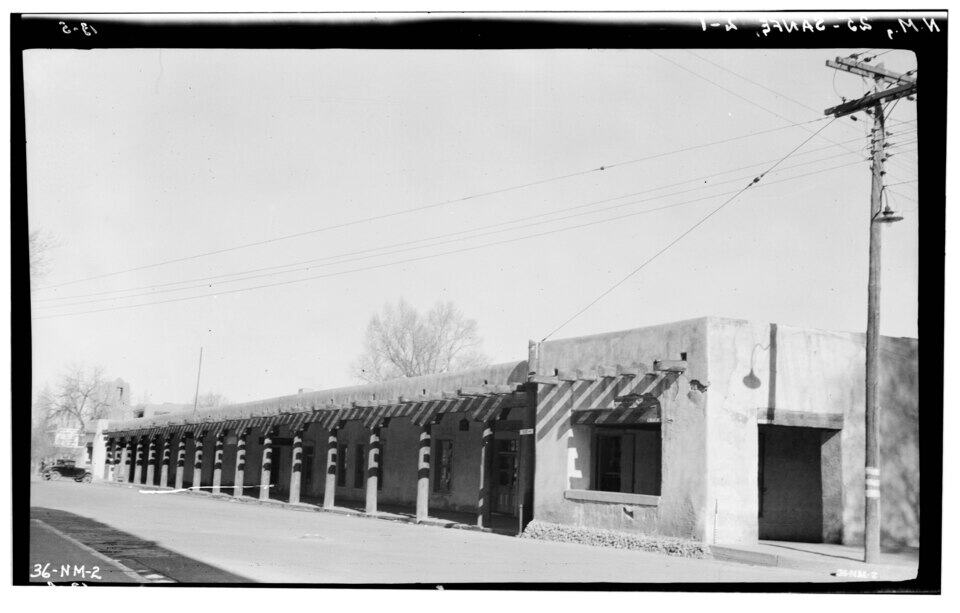 A black-and-white photograph of a one story adobe building with a long covered patio. 