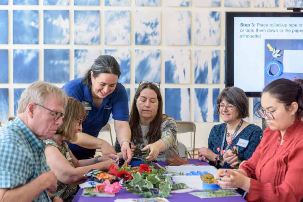 People sit and kneel surround a table filled with art supplies. A blue and white mural and large screen is in the background.
