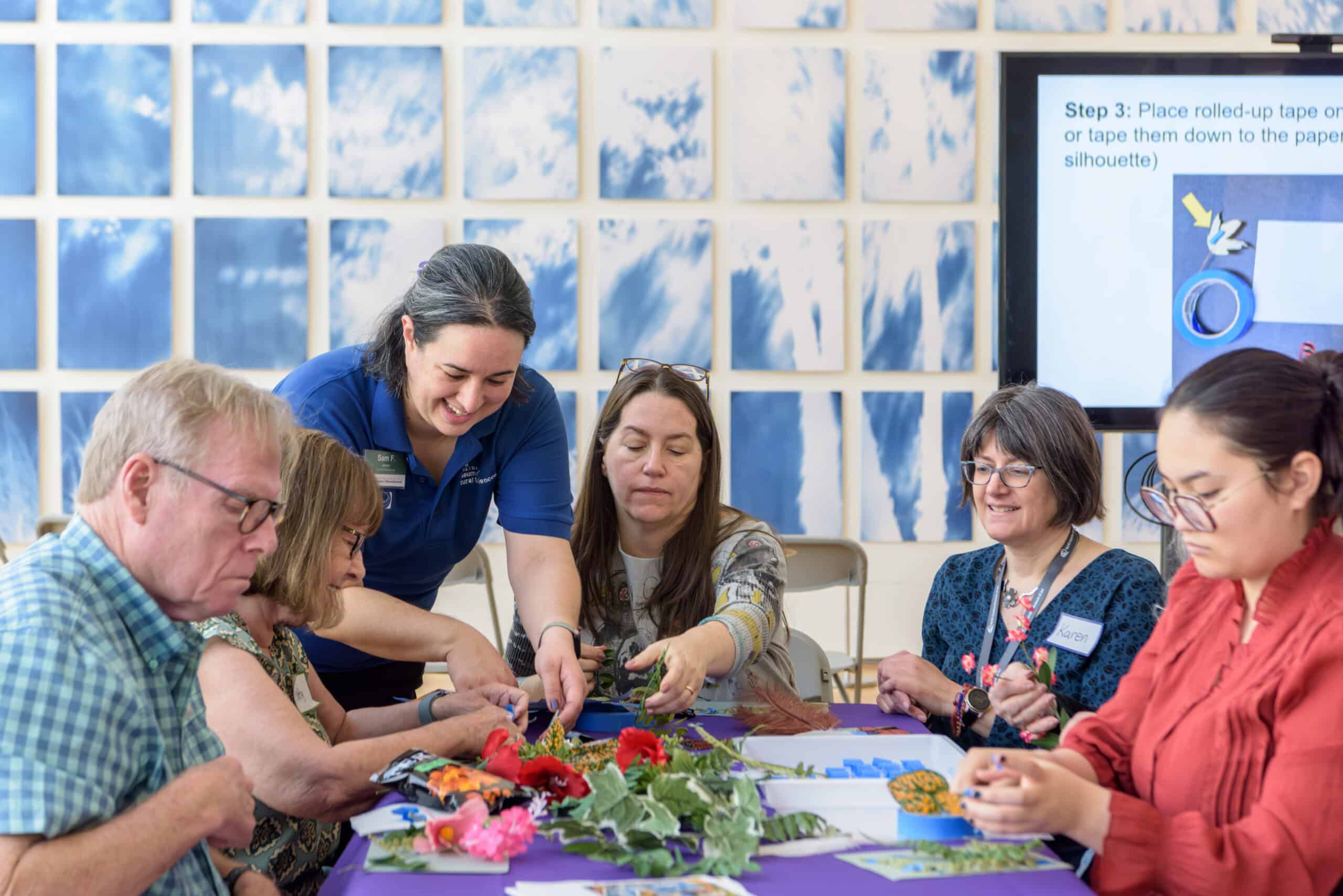 People sit and kneel surround a table filled with art supplies. A blue and white mural and large screen is in the background.