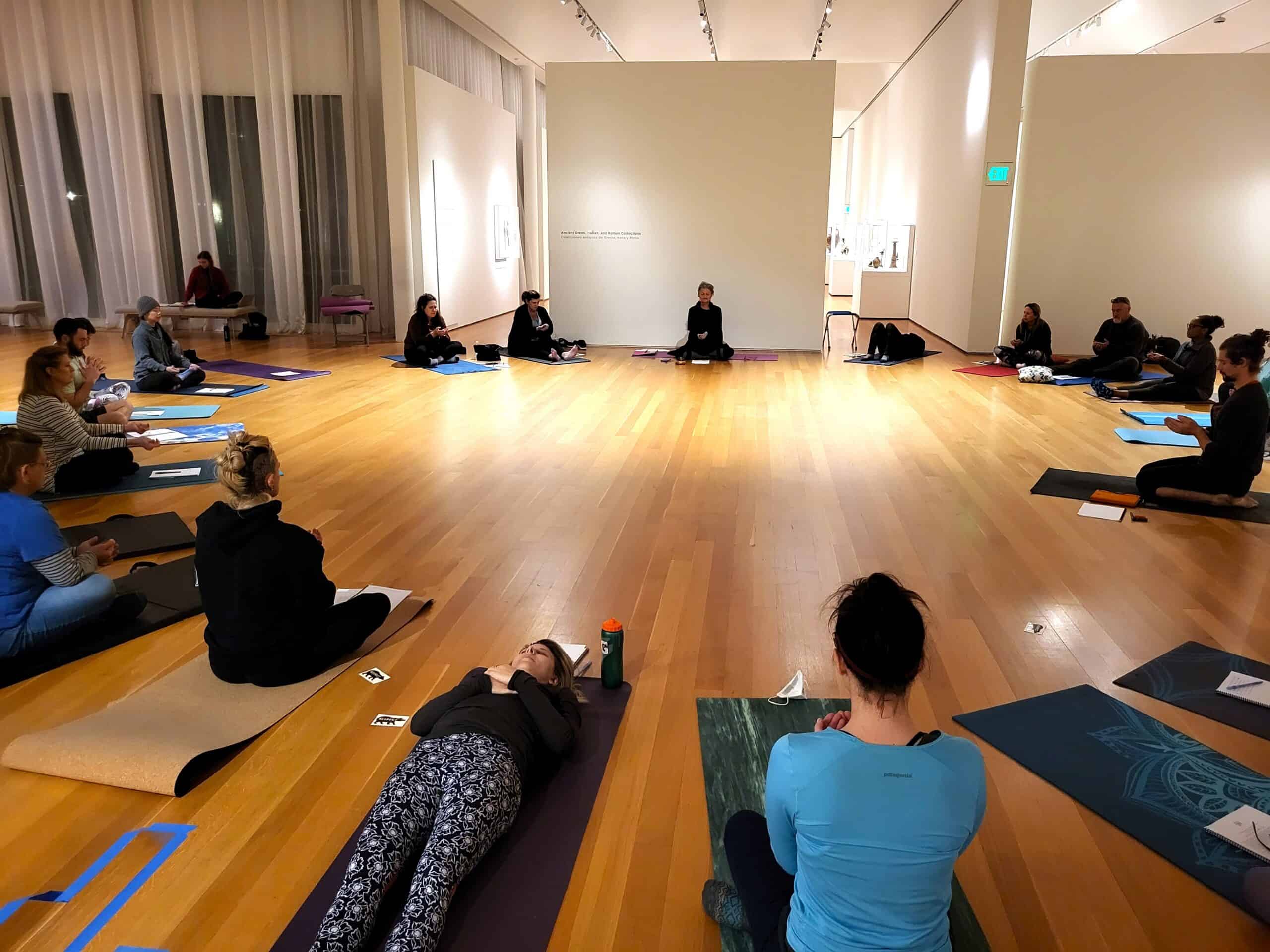 People sit on mats on the floor in the shape of circle in a large open gallery.