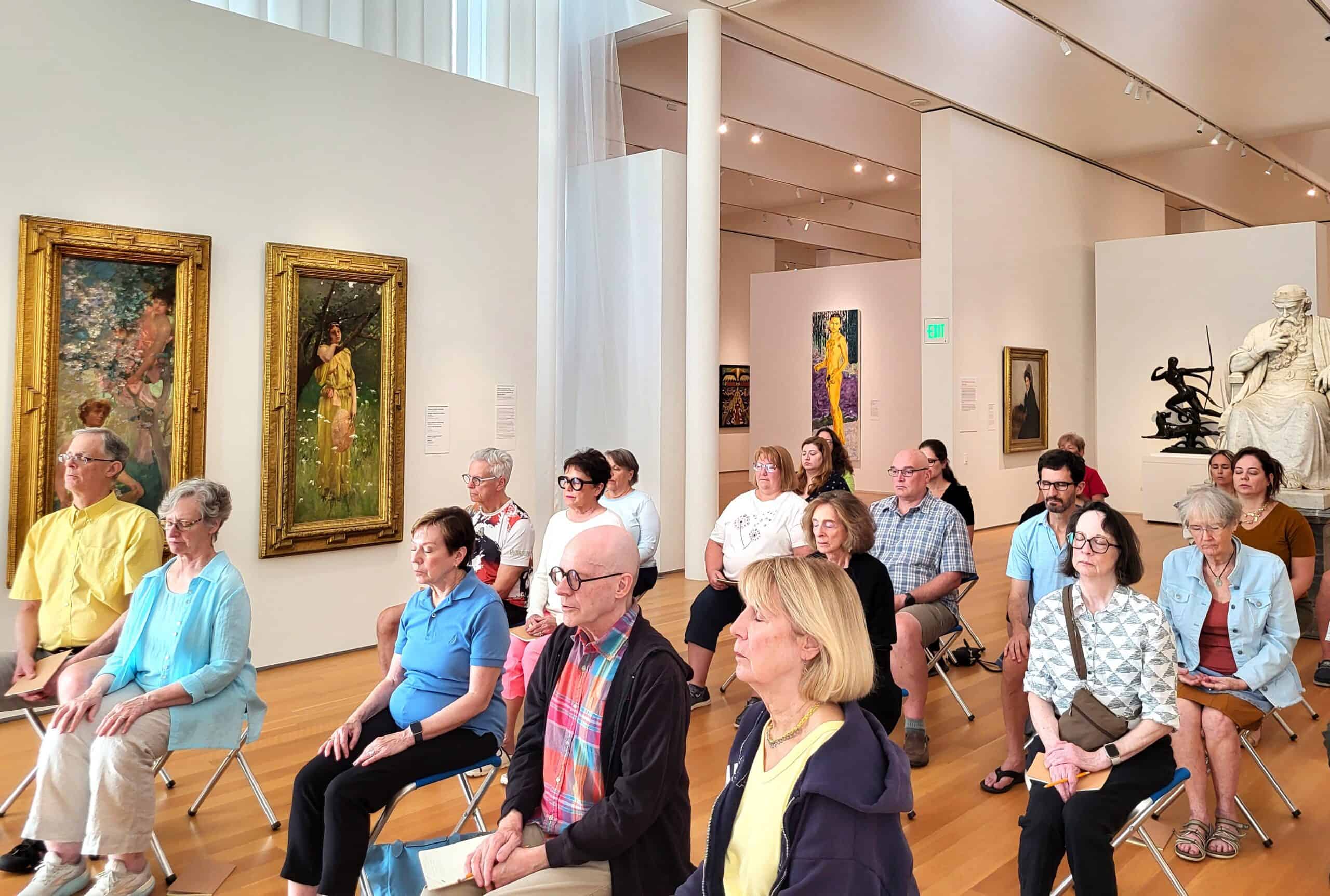 Twenty people with their eyes closed sit on small stools in a bright gallery surrounded by paintings and sculpture.