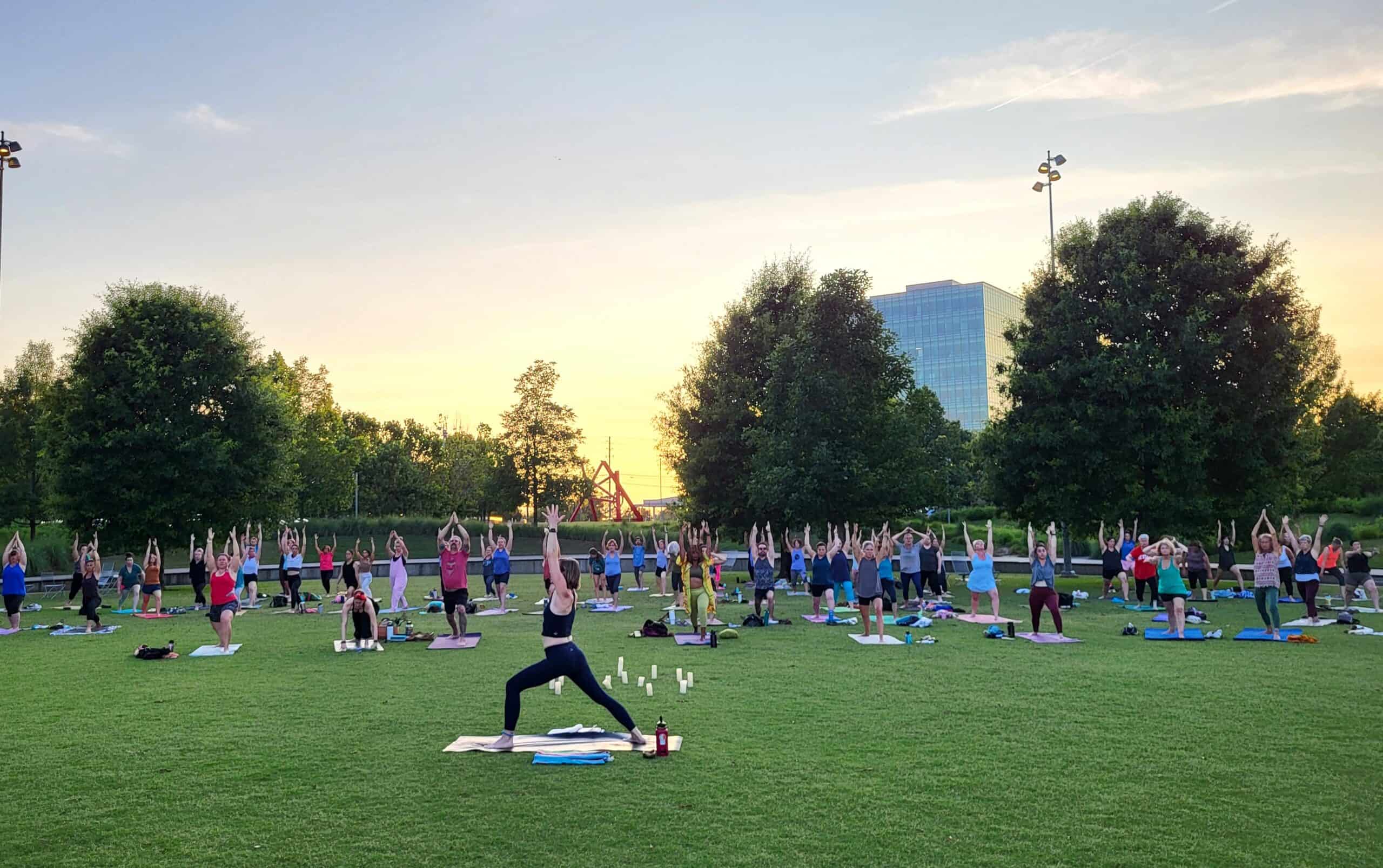 Large group of people on yoga mats in a grassy field hold a standing yoga pose with the sun setting behind them.