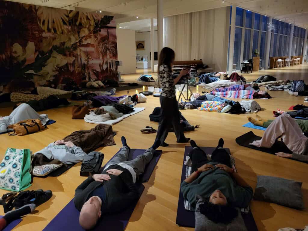 A group of people lie on mats in a large circle on the floor of an art gallery while a woman walks among them.