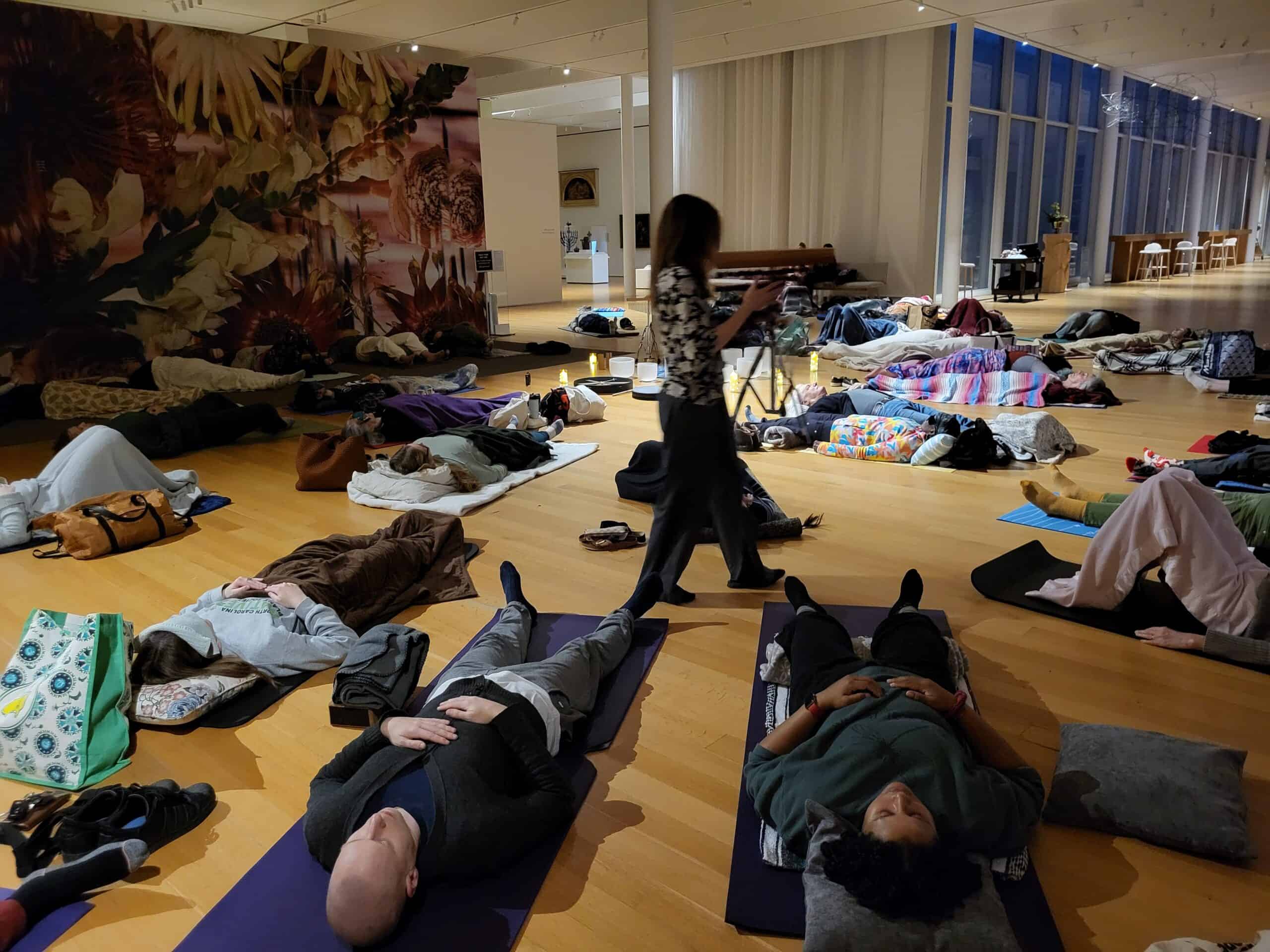 A group of people lie on mats in a large circle on the floor of an art gallery while a woman walks among them.