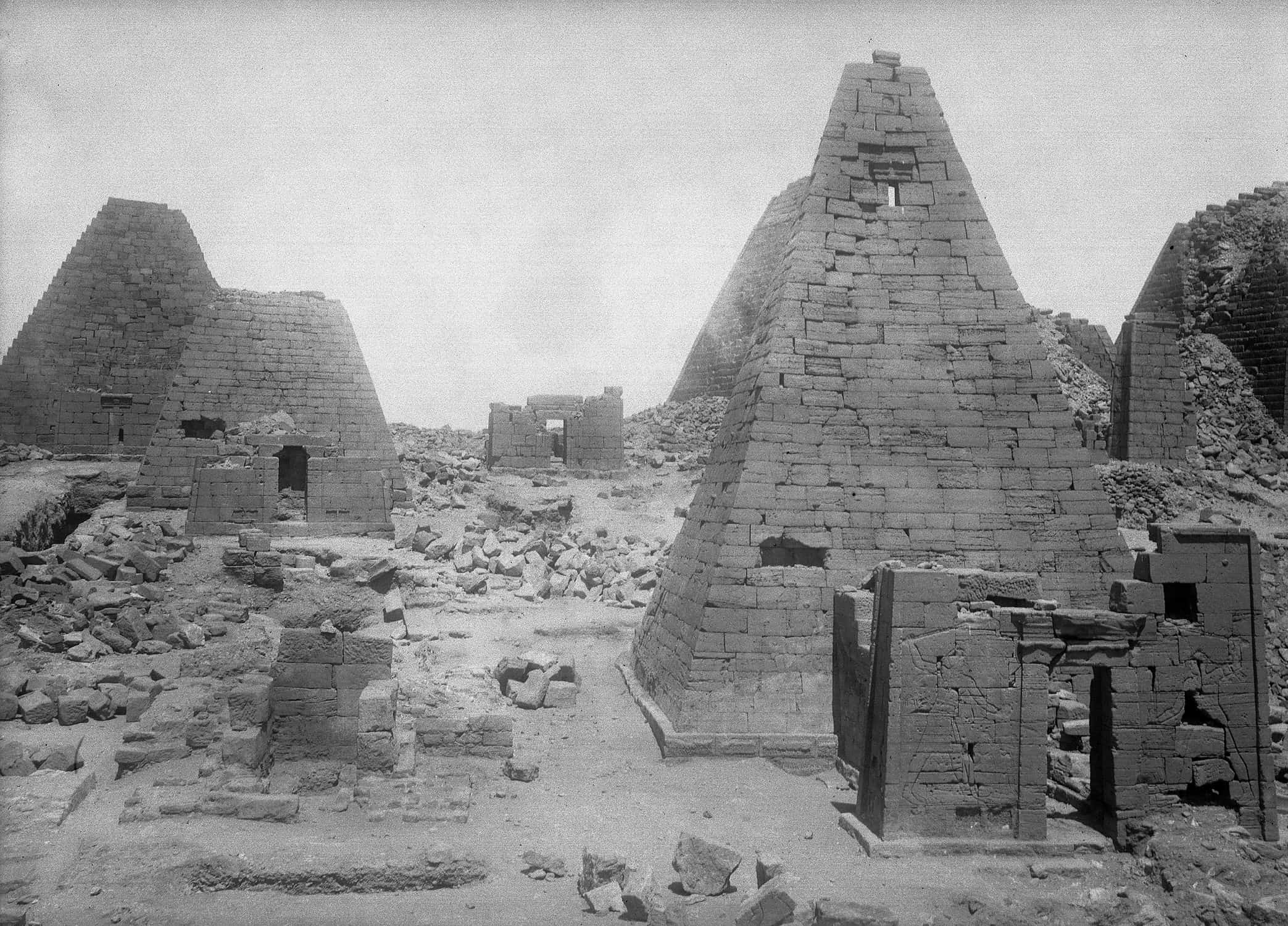A black-and-white photograph of a complex of architectural ruins. Five or six pyramids are in various states of deterioration.