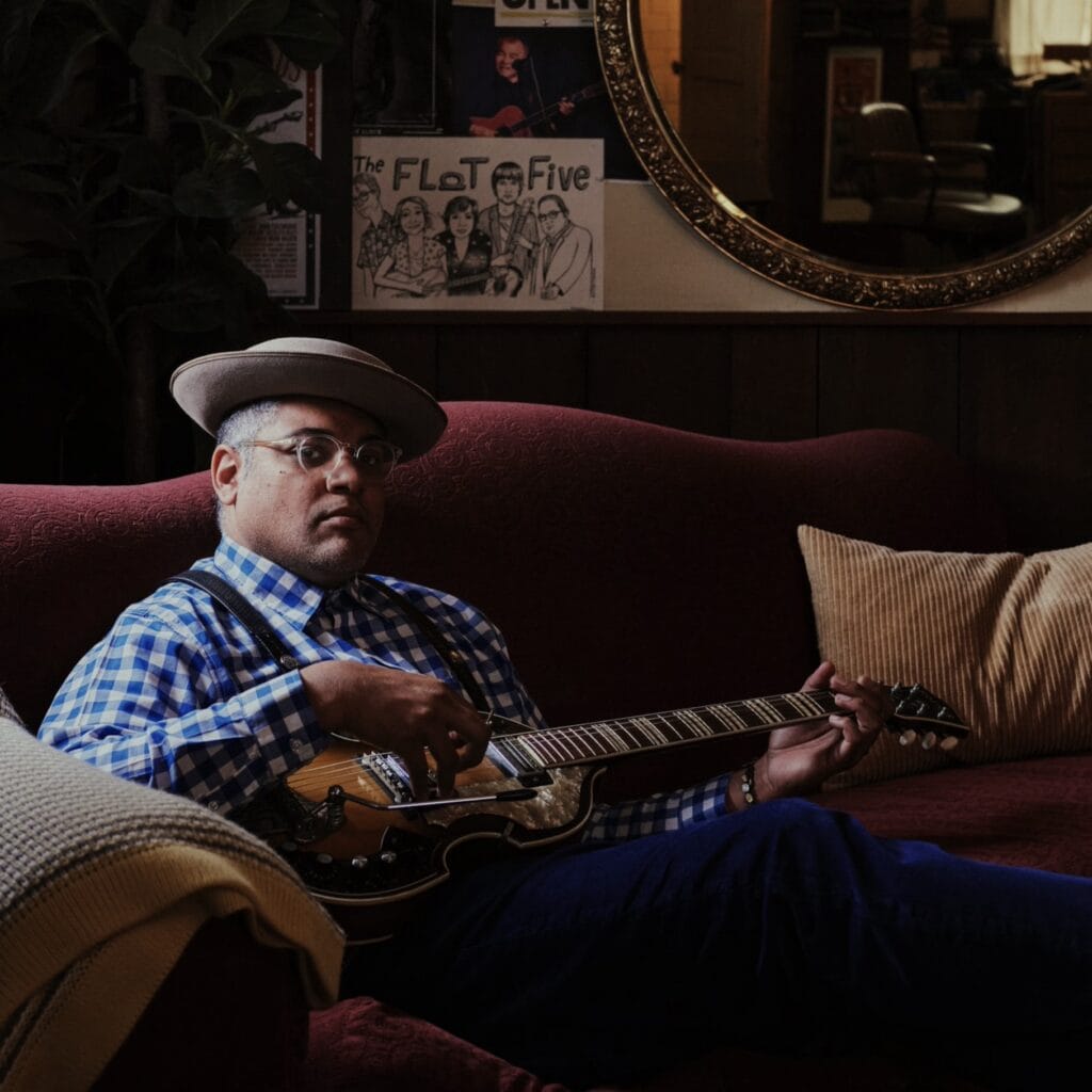 A man in a hat playing a guitar sits on a couch in a darkened room and looks out at the viewer.