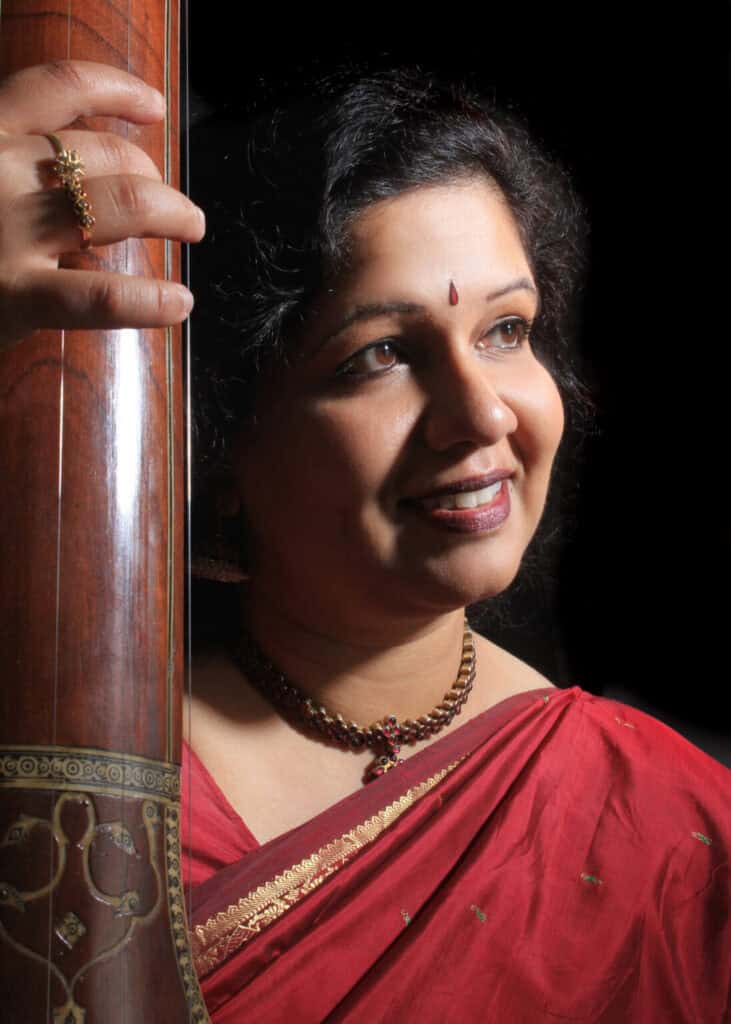 Bust-length photographic portrait of a woman in a red sari holding the neck of a stringed instrument.