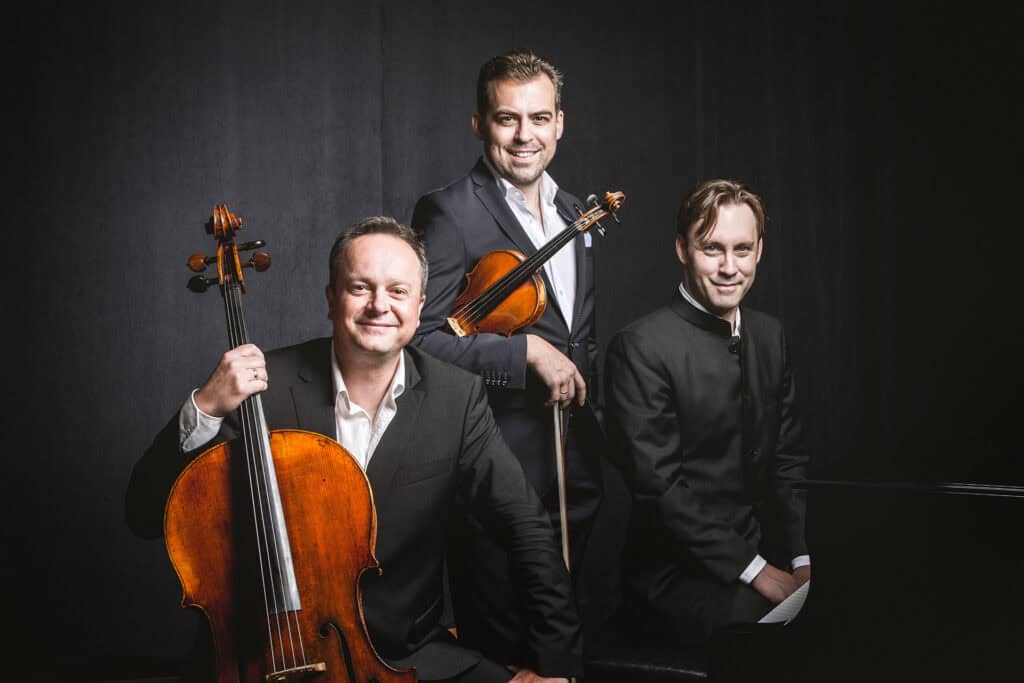 Members of the Phaeton Piano Trio sit and stand next to a piano. Two of the men hold stringed instruments.