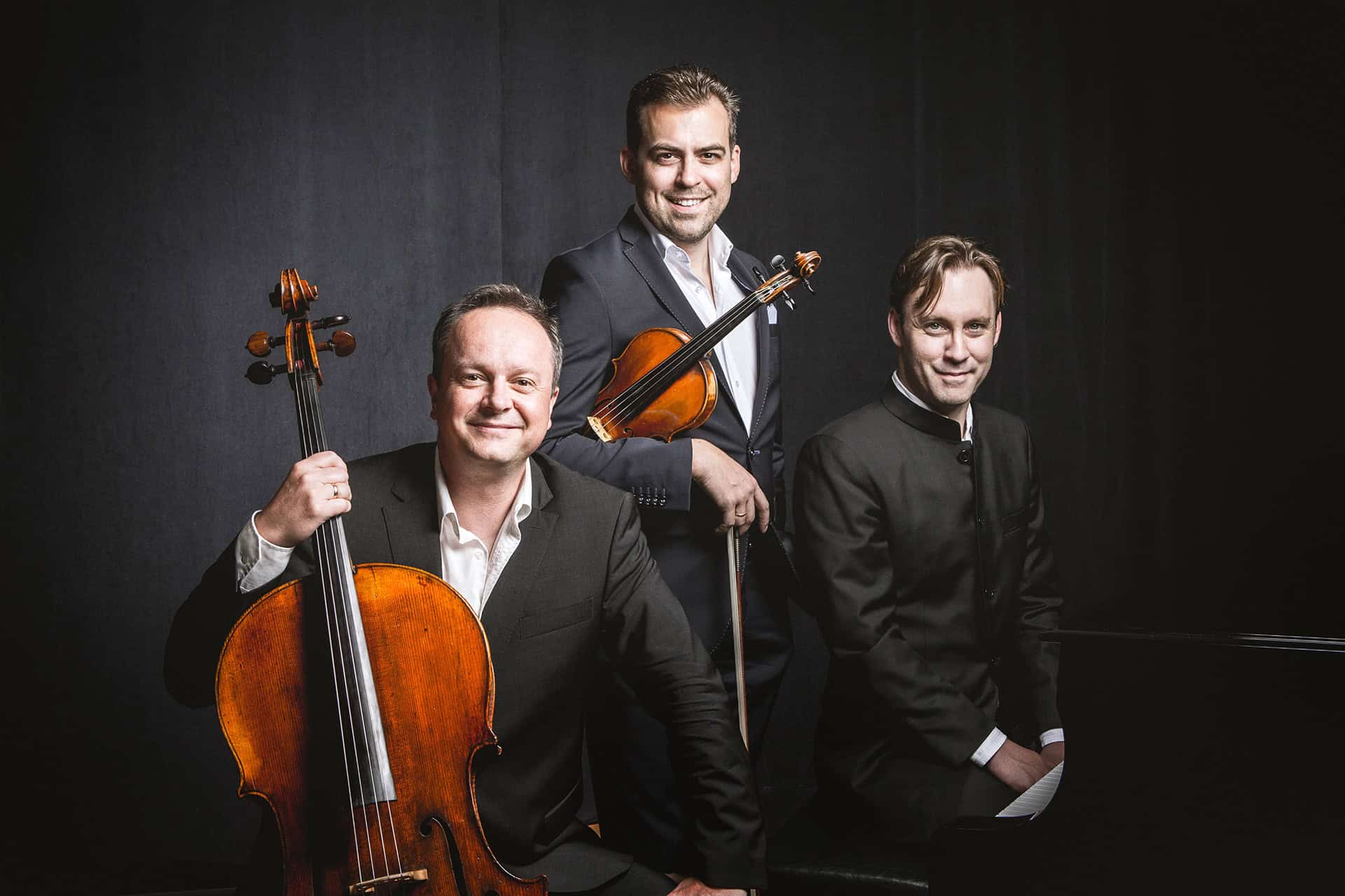 Members of the Phaeton Piano Trio sit and stand next to a piano. Two of the men hold stringed instruments.