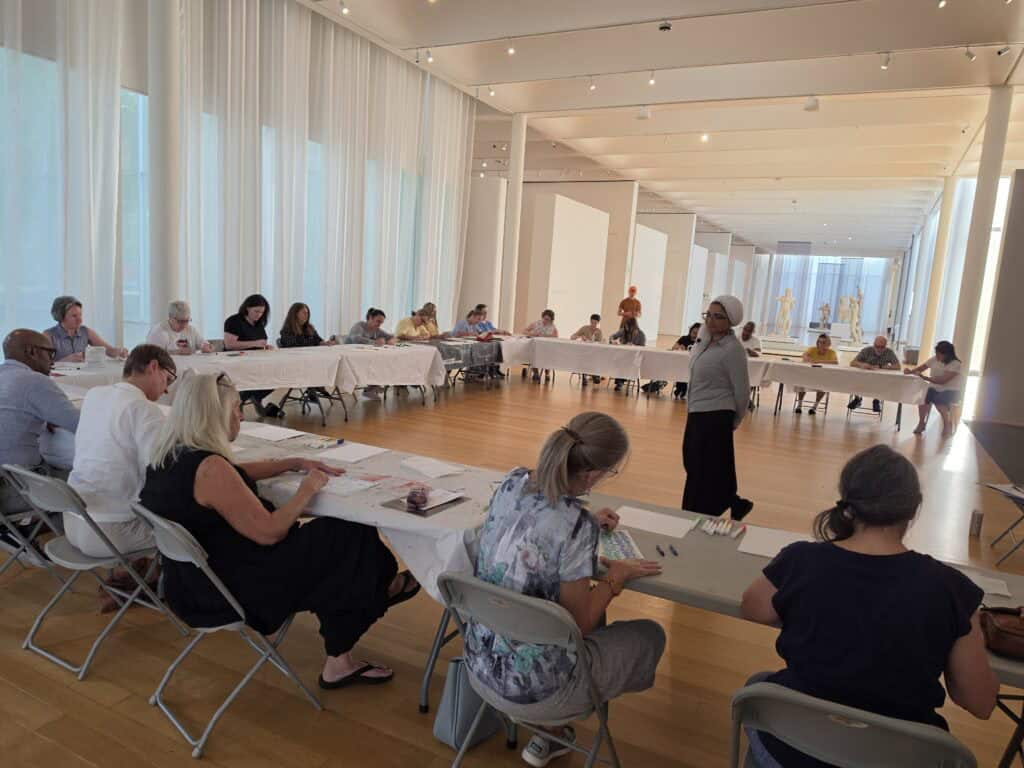 People sit at tables and draw on paper in a large open space in an art gallery.