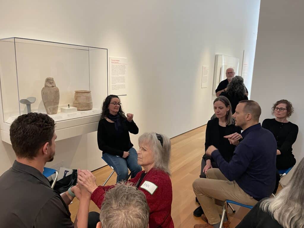 A docent signs in front of ancient Egyptian objects with a group of tour participants sitting in front of her.