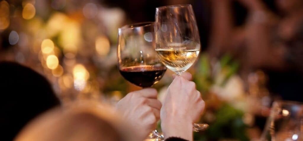 Two hands clink glasses of red and white wine with warm candlelight and a softly blurred table setting in the background.