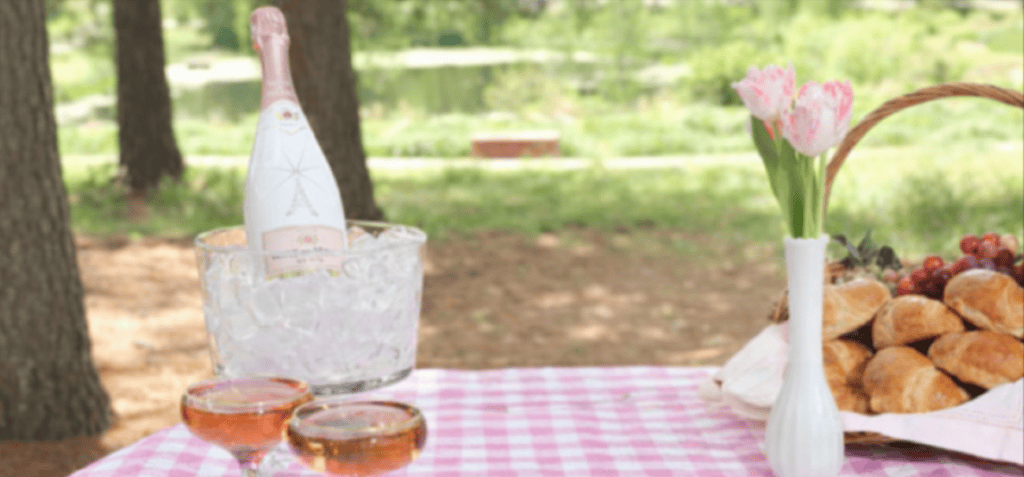 An ice bucket with a bottle of wine, two glasses, a vase with tulips, and a basket of croissants and grapes on a picnic table.