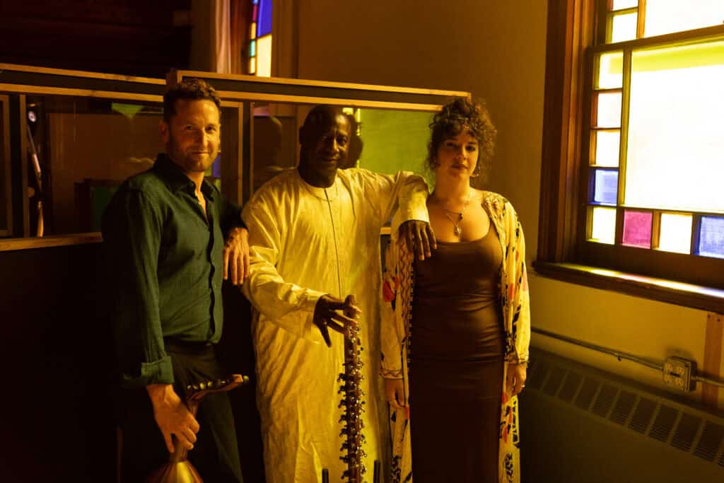 Two men and one woman stand at the back of a row of pews next to a stained glass window. Each man holds a large stringed instrument.