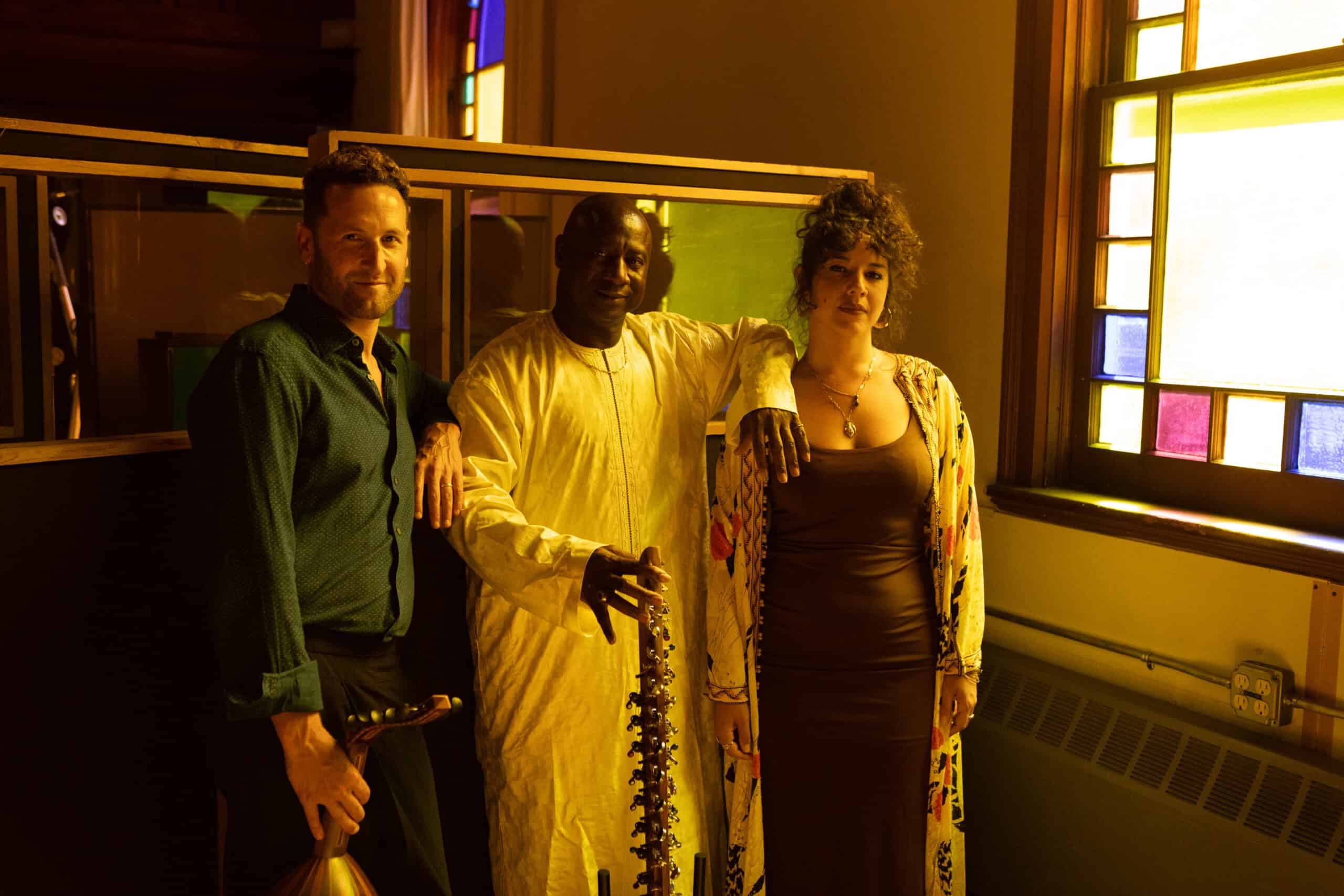 Two men and one woman stand at the back of a row of pews next to a stained glass window. Each man holds a large stringed instrument.