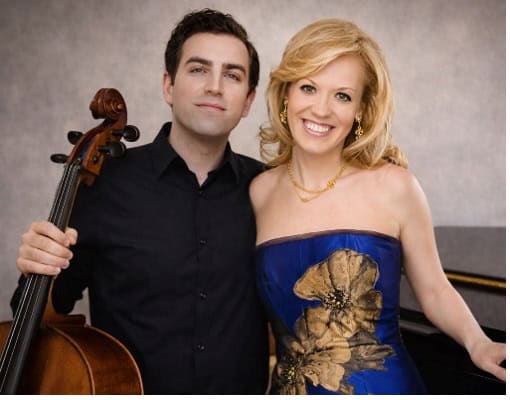 A man in a black shirt holding the neck of a cello stands next to a woman in a blue evening gown leaning on a piano.