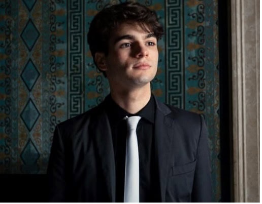 A young man in a black shirt and coat and white tie gazes off to the right in front of a wall with geometric patterns.