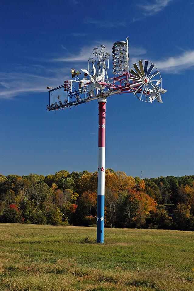 Metal sculpture made of spare parts painted red, white, and blue at the top of a tall pole in the middle of a field.