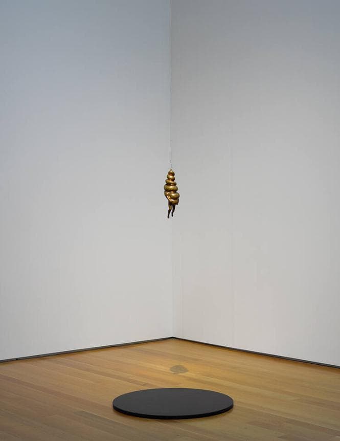 Sculpture of a women whose torso and head are a bulbous spiral suspended by a wire over a dark slate circle placed on the floor.