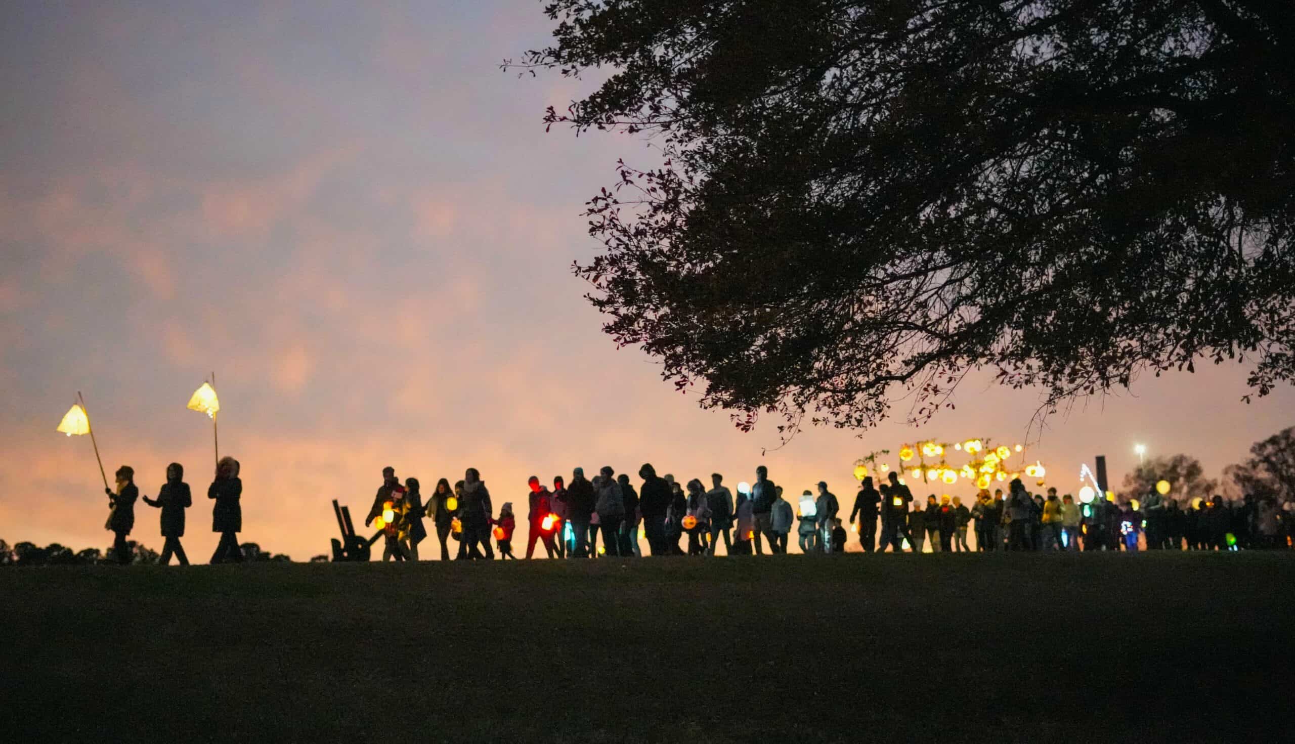 A large group of people seen from a distance walk and carry lanterns of all shapes and sizes in a landscape at dusk.