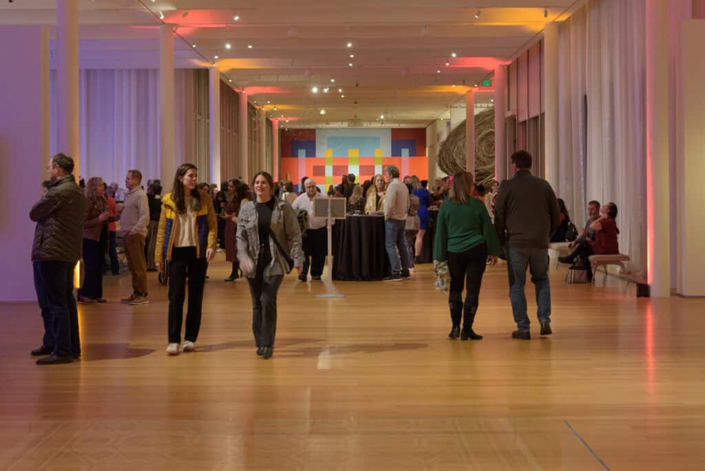 A crowd of people walk around and mingle in a colorfully lit art gallery setting.