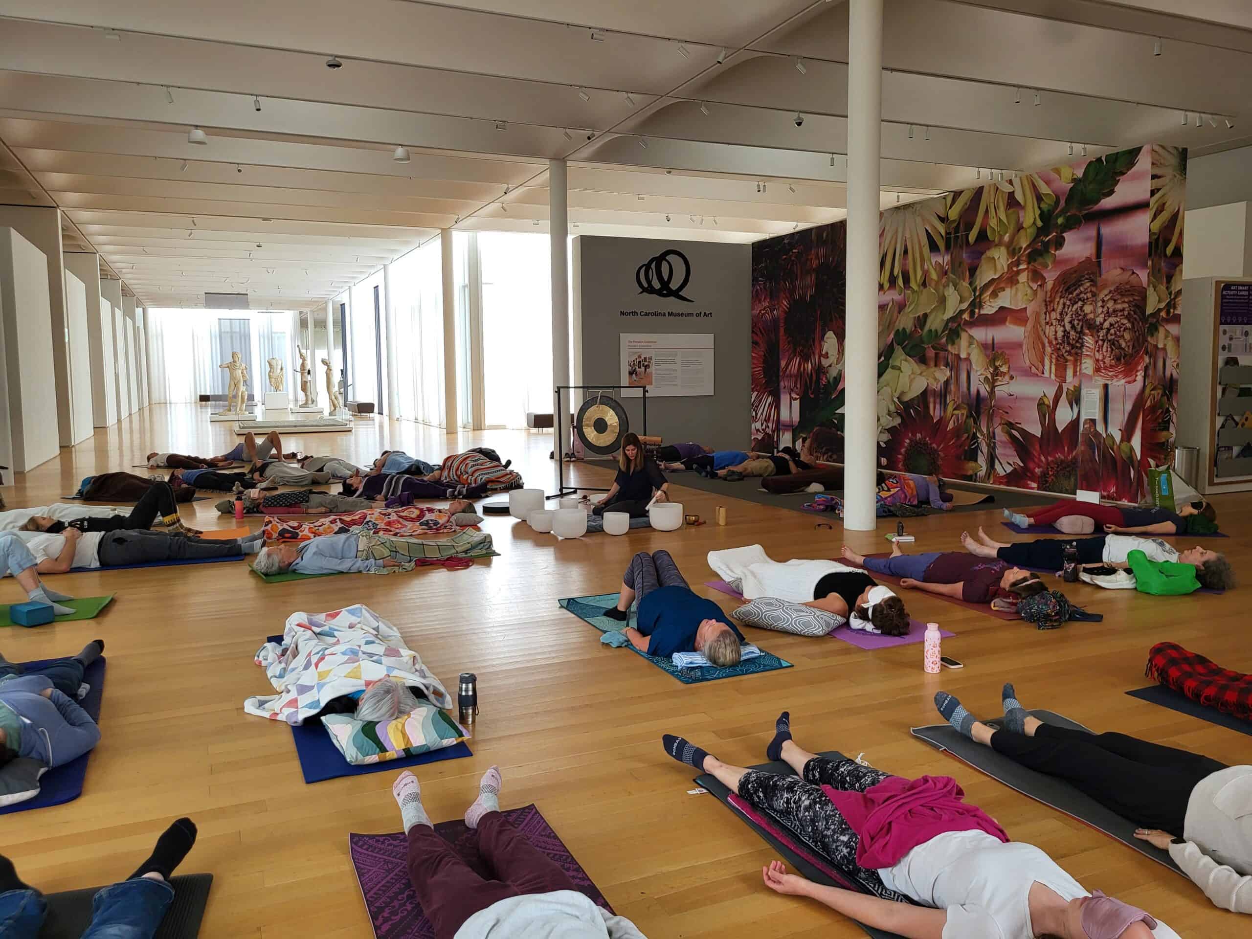 A group of people lie on mats on the floor of an art gallery while a woman plays crystal singing bowls in the center.