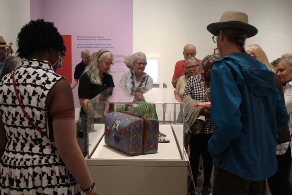 A group of people stand around a display case examining two beaded suitcases in an art gallery.