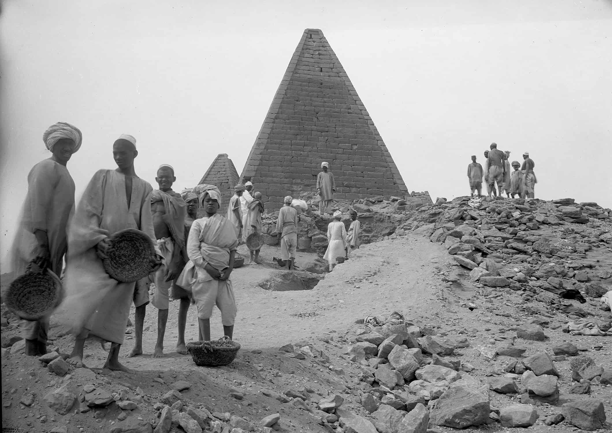 A black-and-white photograph of several groups of men standing near a path of rubble with two pyramids in the background.