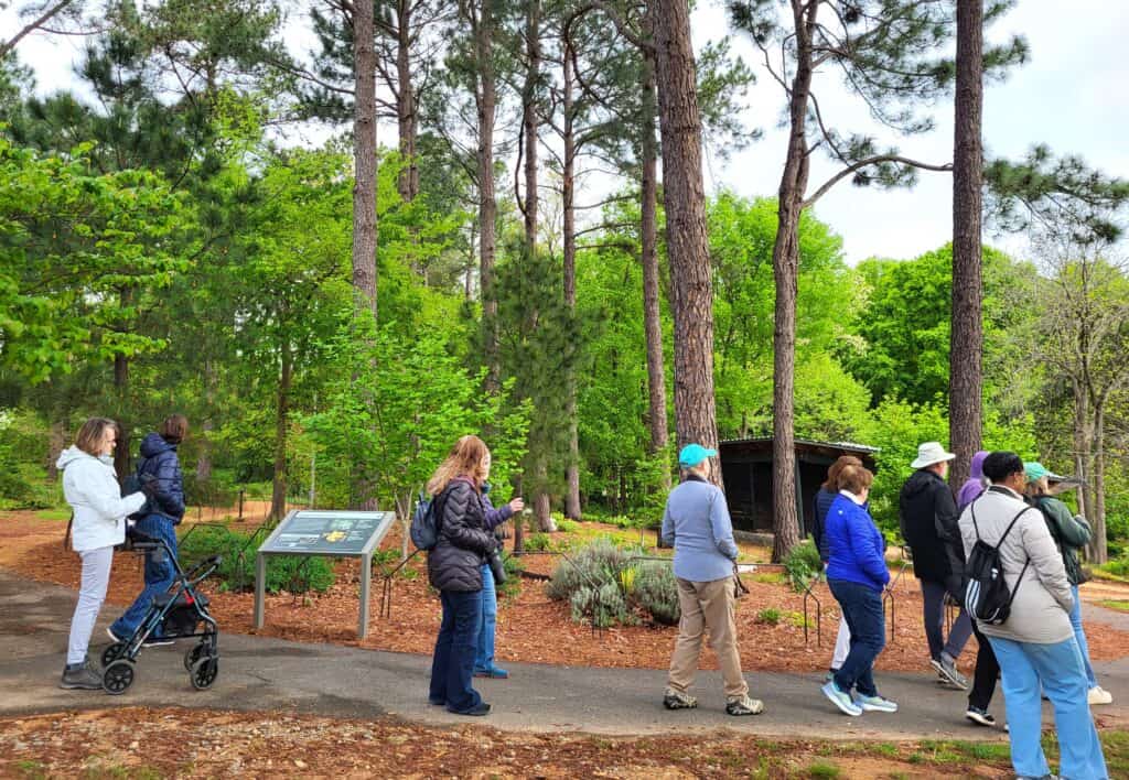 A group of people dressed for the outdoors walk on a paved path surrounded by grass, trees, and flowering plants.