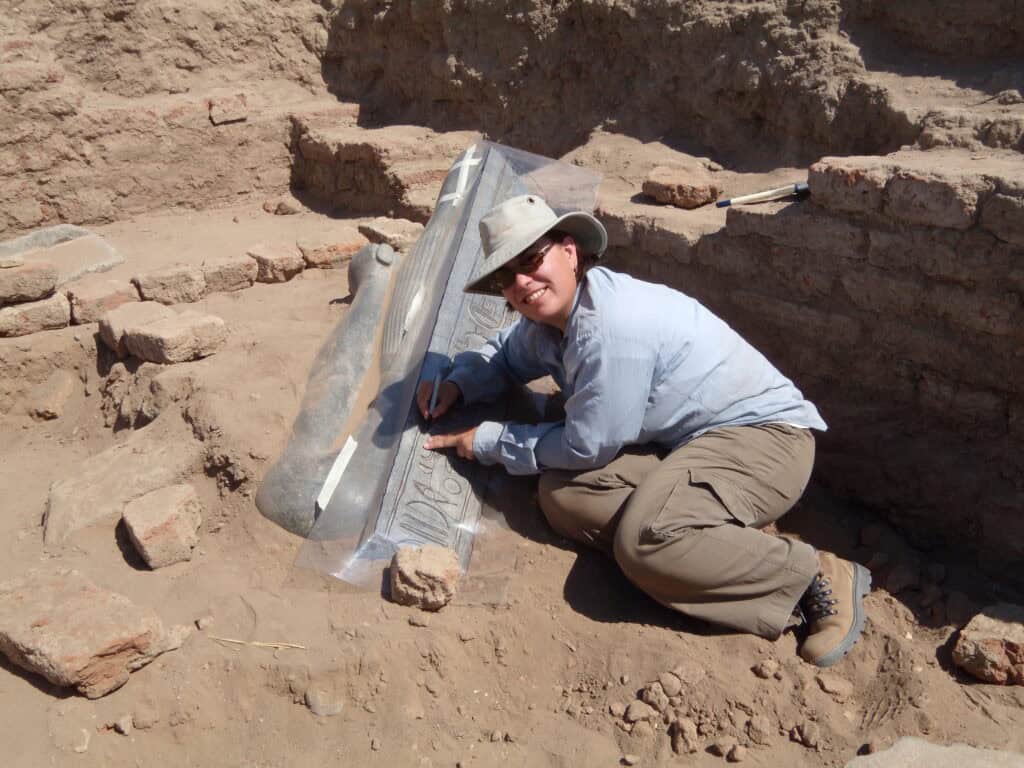A woman near a crumbling brick wall kneels and traces the text on a partially buried object covered with a sheet of plastic.