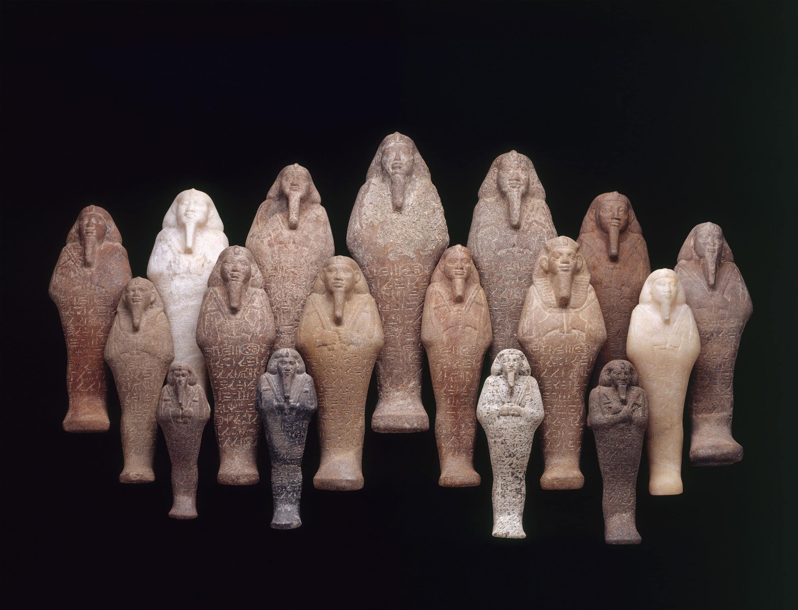 Frontal view of 17 stylized figures of various sizes with headdresses, long, narrow beards, and writing on their torsos and legs.