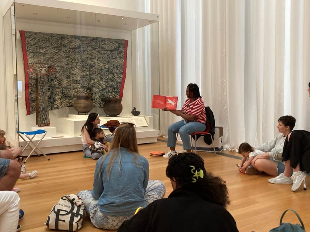 A woman reads a book to a group of children and adults seated on the floor in a museum gallery with pottery and textile displays.