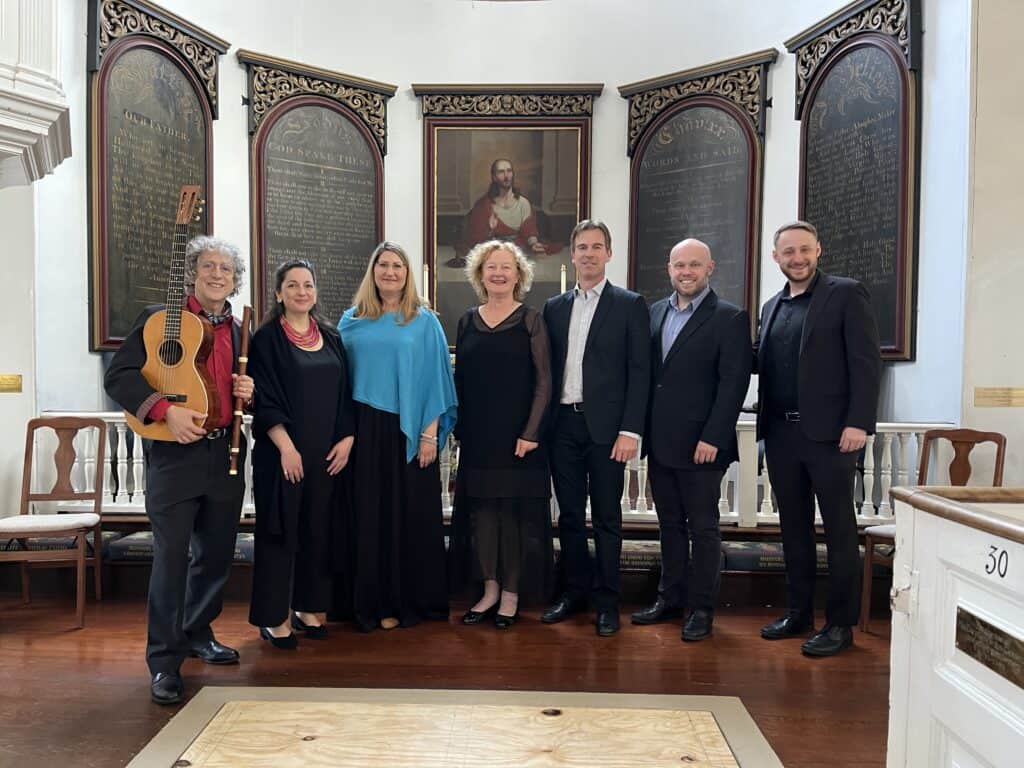 Seven people, four men and three women, stand side by side in a church. The first man on the left holds a guitar and wind instrument.]