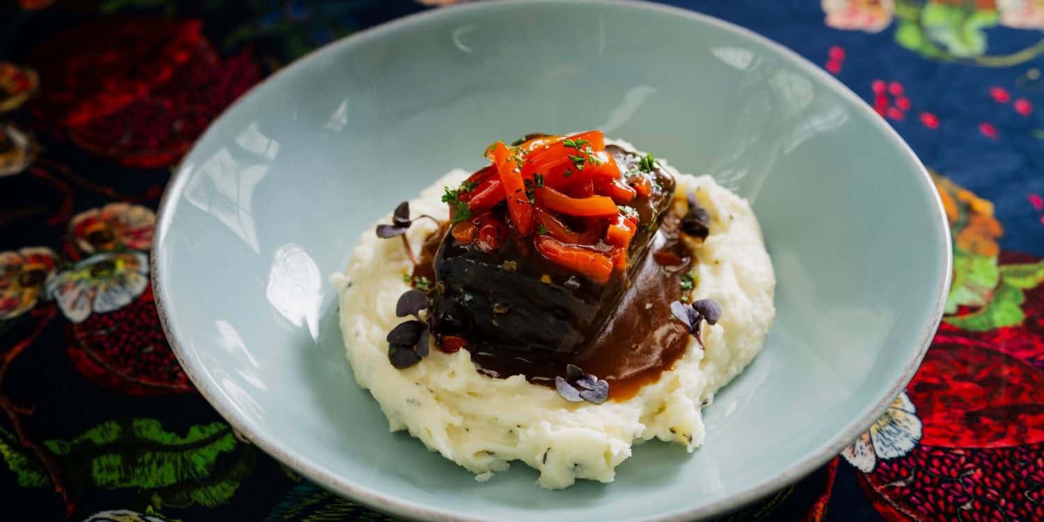 A plated entrée featuring a beef short rib over a bed of mashed potatoes with a brown sauce on a colorful floral tablecloth.