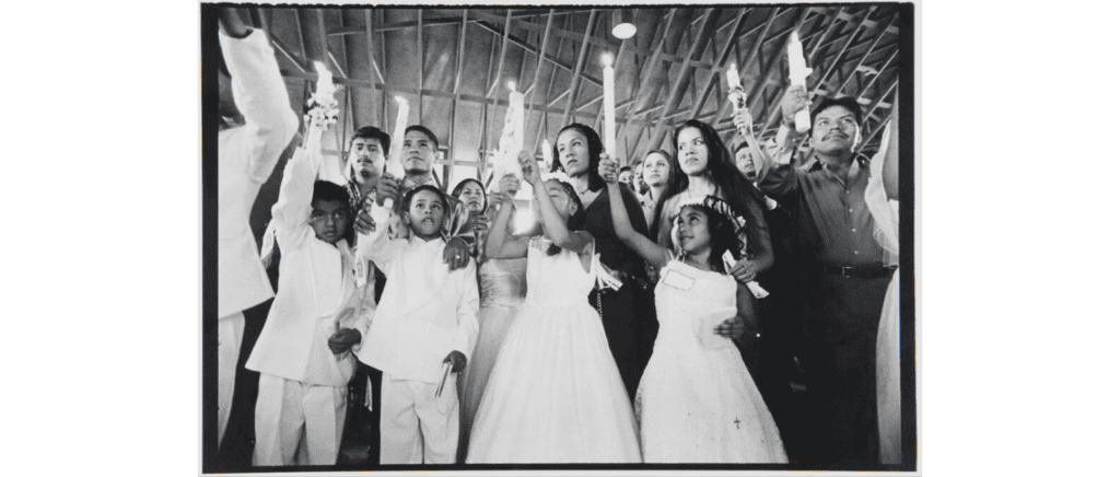 A group of children dressed in white dresses and suits as well as the adults behind them raise lit candles in a large indoor space.