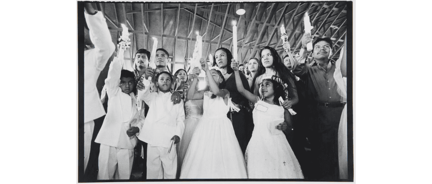 A group of children dressed in white dresses and suits as well as the adults behind them raise lit candles in a large indoor space.