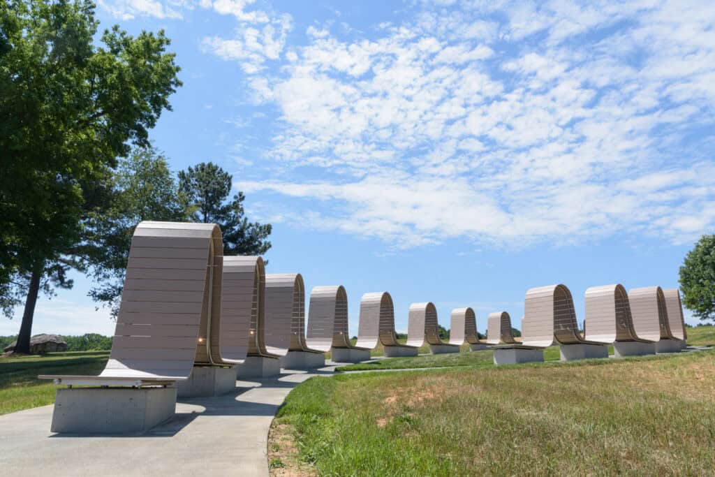 Artwork with an undulating form made of tan decking on a steel frame above concrete blocks, flanked by tall trees in a park.
