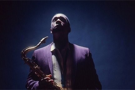 John Coltrane, lit from above, holds his saxophone and looks up.