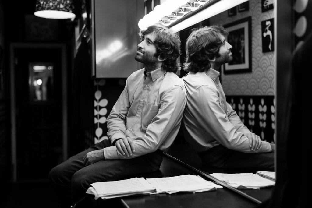 A man sits on a table and leans on a large mirror in a dressing room.