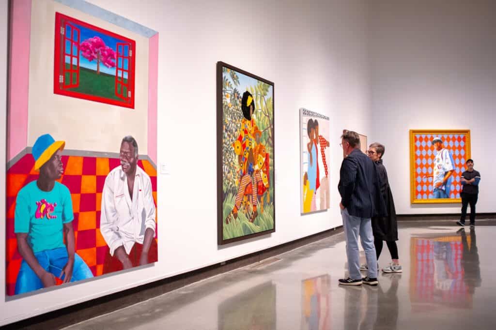 Three people view large, colorful paintings in an white-walled art gallery with a high ceiling.