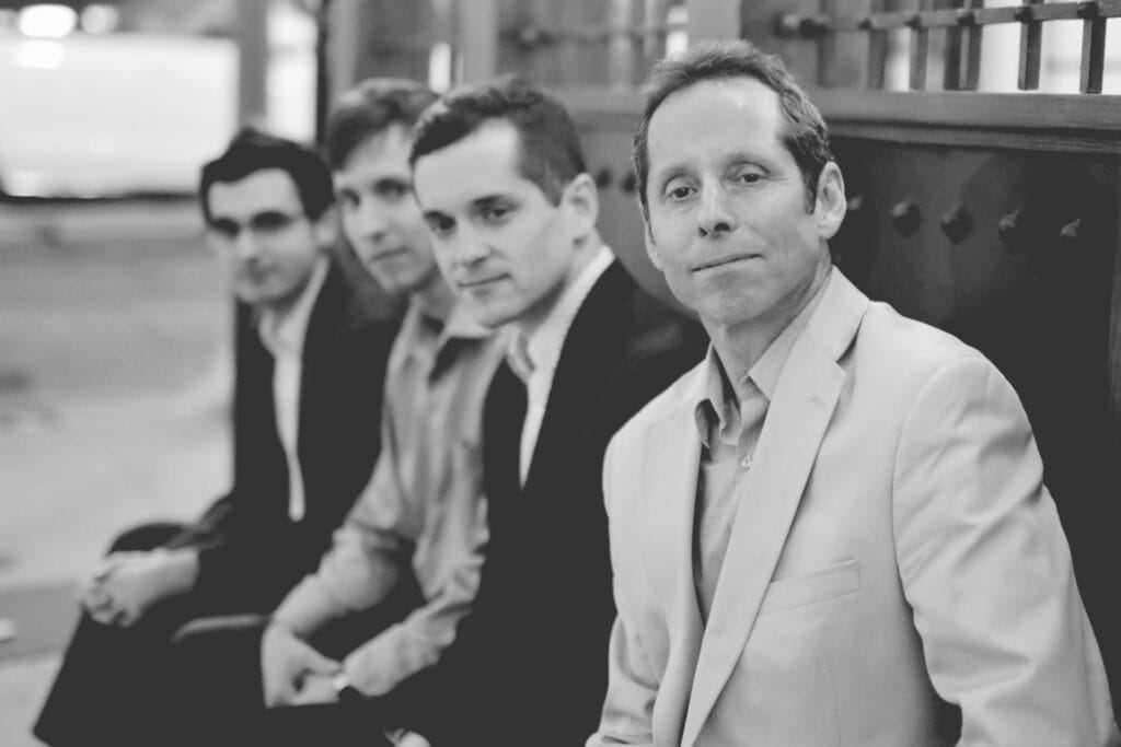 Four men in suit jackets sit on a high-back bench placed on a diagonal to the picture plane.