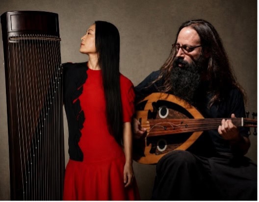 A standing woman holds up and looks at a Chinese plucked zither while a seated man looks down at and plays an oud.