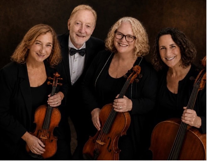 Three women and one man in formal attire look out at the viewer and smile. The woman each hold a stringed instrument.