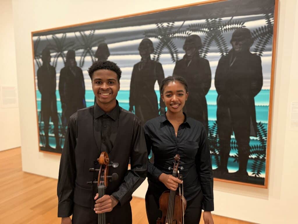 Two youth performers stand holding a cello and violin in front of an artwork in an art gallery.