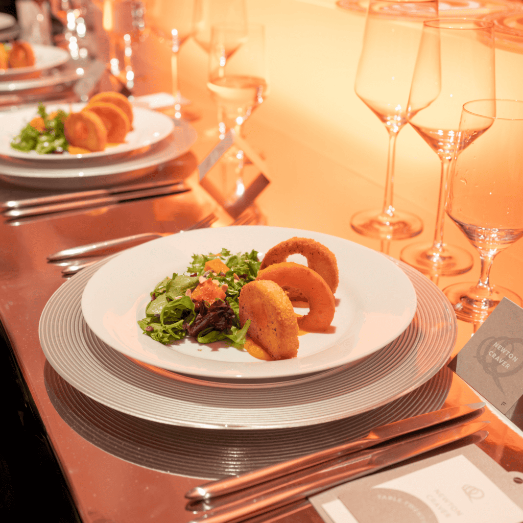 Photograph of an elegant restaurant table set for fine dining, featuring a white plate with plated salad, elegant ring striped chargers, silverware, wine glasses, and warm candlelight.